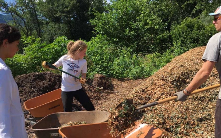 Two people shovel mulch into a wheelbarrow, held by another. 